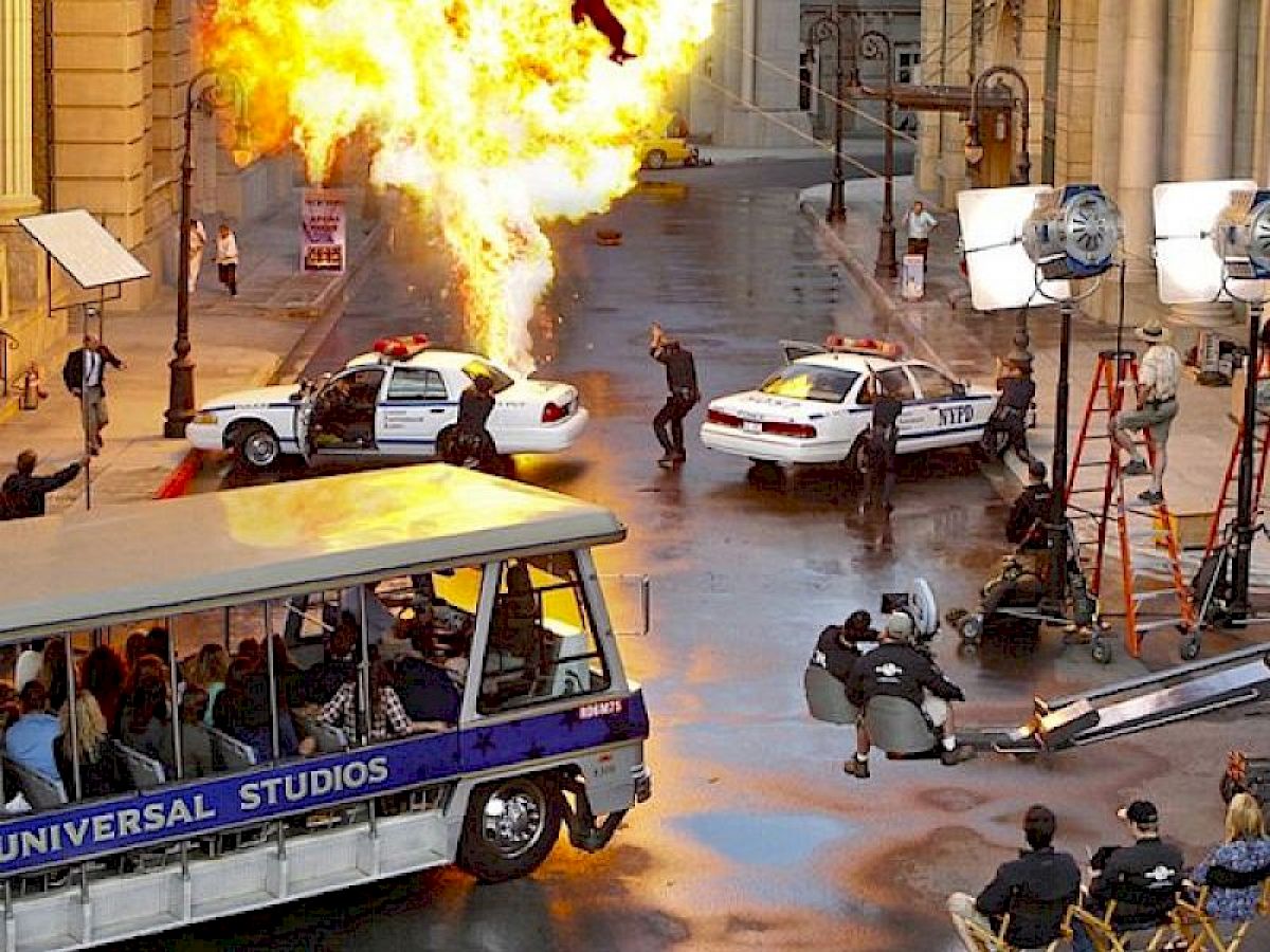 A movie set with an explosion, police cars, crew filming the action, and a tram tour labeled "Universal Studios" nearby, ending the sentence.