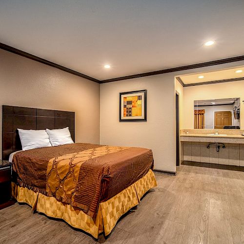 A cozy hotel room with a single bed, bedside table, framed artwork, and an open area leading to a bathroom counter with a sink and mirror.