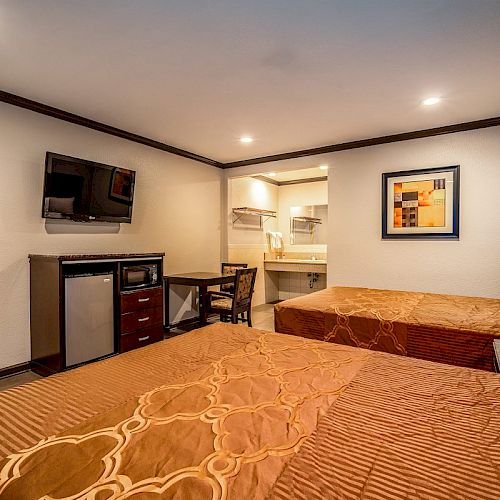 A hotel room with two beds, a wall-mounted TV, a mini-fridge, a microwave, a desk, and a chair, featuring a framed artwork above a headboard.