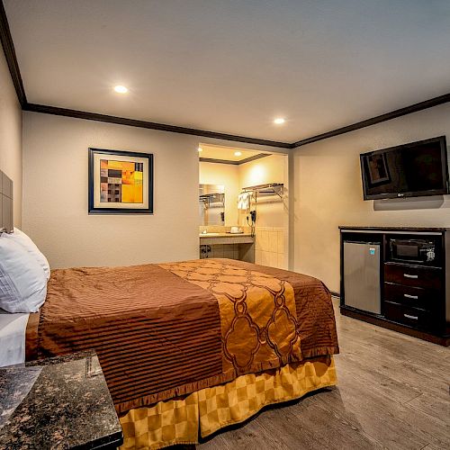 This image shows a small hotel room with a bed, a wall-mounted TV, a mini-fridge, a microwave, and a piece of art above the bed's headboard.