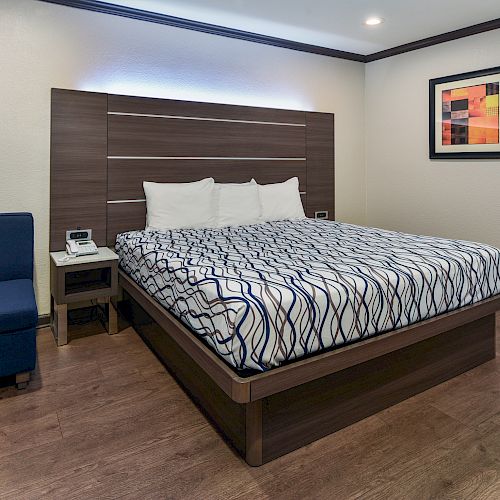 A modern hotel room features a bed with patterned bedding, a navy blue armchair, a nightstand, and a framed abstract painting on the wall.