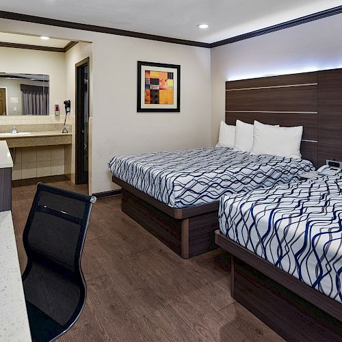 This image shows a hotel room with two double beds, a desk and chair, a sink area, and modern decor. The walls are light-colored and there is a framed artwork.