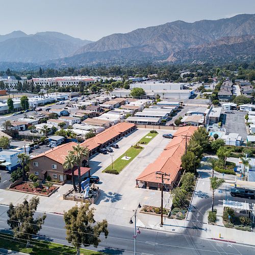 The image shows an aerial view of a suburban area with residential buildings, streets, trees, and mountains in the background, taken during the daytime.