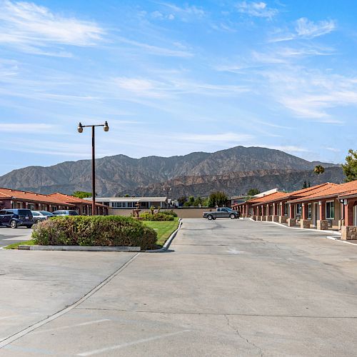 The image shows a motel with parking spaces, vehicles, and one-story buildings with a mountain backdrop under a blue sky.