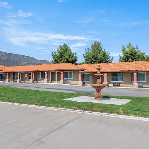 A single-story motel with a red-tiled roof, neat landscaping, and a central fountain, set against a backdrop of trees and mountains.