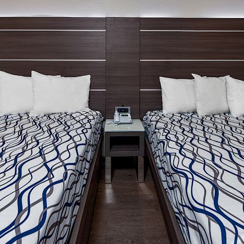 The image features two beds with patterned bedspreads, white pillows, and a wooden headboard, separated by a nightstand holding a digital clock.