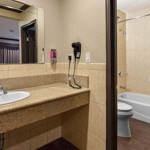 The image shows a bathroom with a sink and countertop, a wall-mounted hairdryer, a bathtub with a shower curtain, and a toilet, all in a tiled setting.