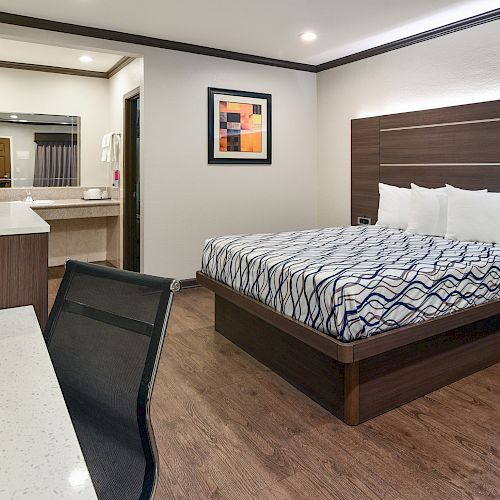 A modern hotel room features a bed, desk, chair, TV, bedside table, and a bathroom area with a mirror and sink in the background.