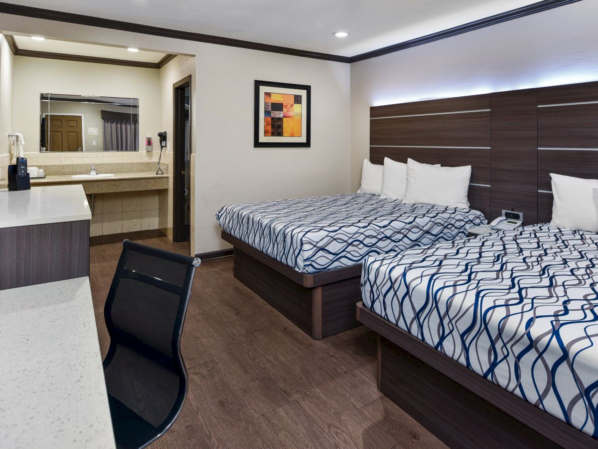 A modern hotel room with two double beds, a desk with an office chair, a wall-mounted TV, a sink area, and framed artwork on the wall.