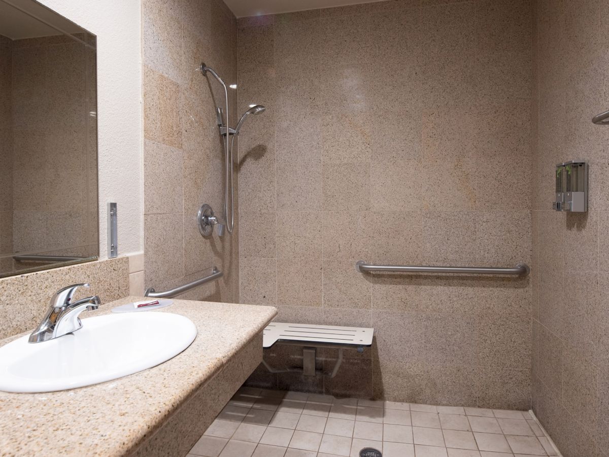 The image shows an accessible bathroom with a sink, shower area with a bench and grab bars, mirror, towel rack, and a wall-mounted soap dispenser.