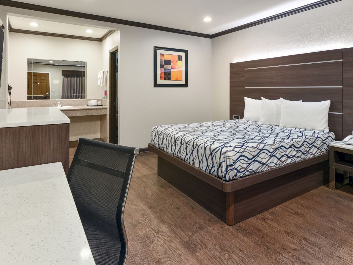 A modern hotel room with a bed, desk, chair, wall art, and a bathroom sink area. The room has wooden floors and contemporary decor.