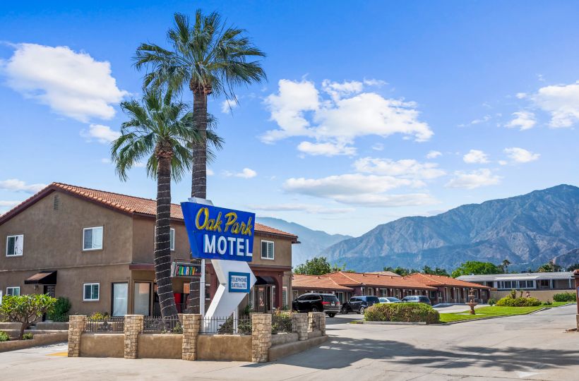 The image shows a motel with a sign reading "Oak Park MOTEL," palm trees, and mountains in the background under a blue sky with clouds.