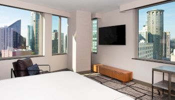 A modern hotel room with cityscape views, a bed, wall-mounted TV, chair, and minimalist furniture, including a small wooden bench.