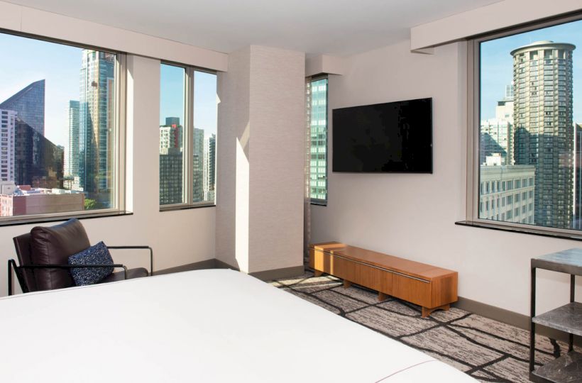 A modern hotel room with cityscape views, a bed, wall-mounted TV, chair, and minimalist furniture, including a small wooden bench.