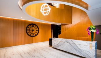A modern lobby with wooden paneling, a large wall clock, a spiral staircase, a marble reception desk, and decorative lighting and flowers.