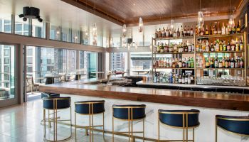 A modern bar with a sleek wooden counter, blue and gold stools, and a fully stocked liquor shelf. Large windows provide a panoramic city view.