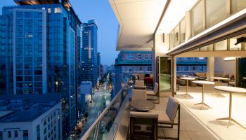 A modern rooftop balcony with seating and tables overlooks a street lined with tall buildings as evening approaches, creating a serene urban view.