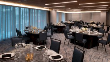 The image shows an elegantly set banquet hall with round tables prepared for a formal event, featuring black napkins, crystal glassware, and floral centerpieces.