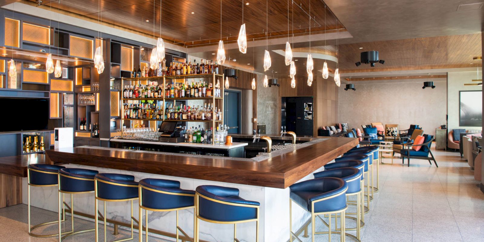 A modern bar with a wooden countertop, high shelves filled with bottles, blue bar stools, elegant lighting, and a seating area in the background.