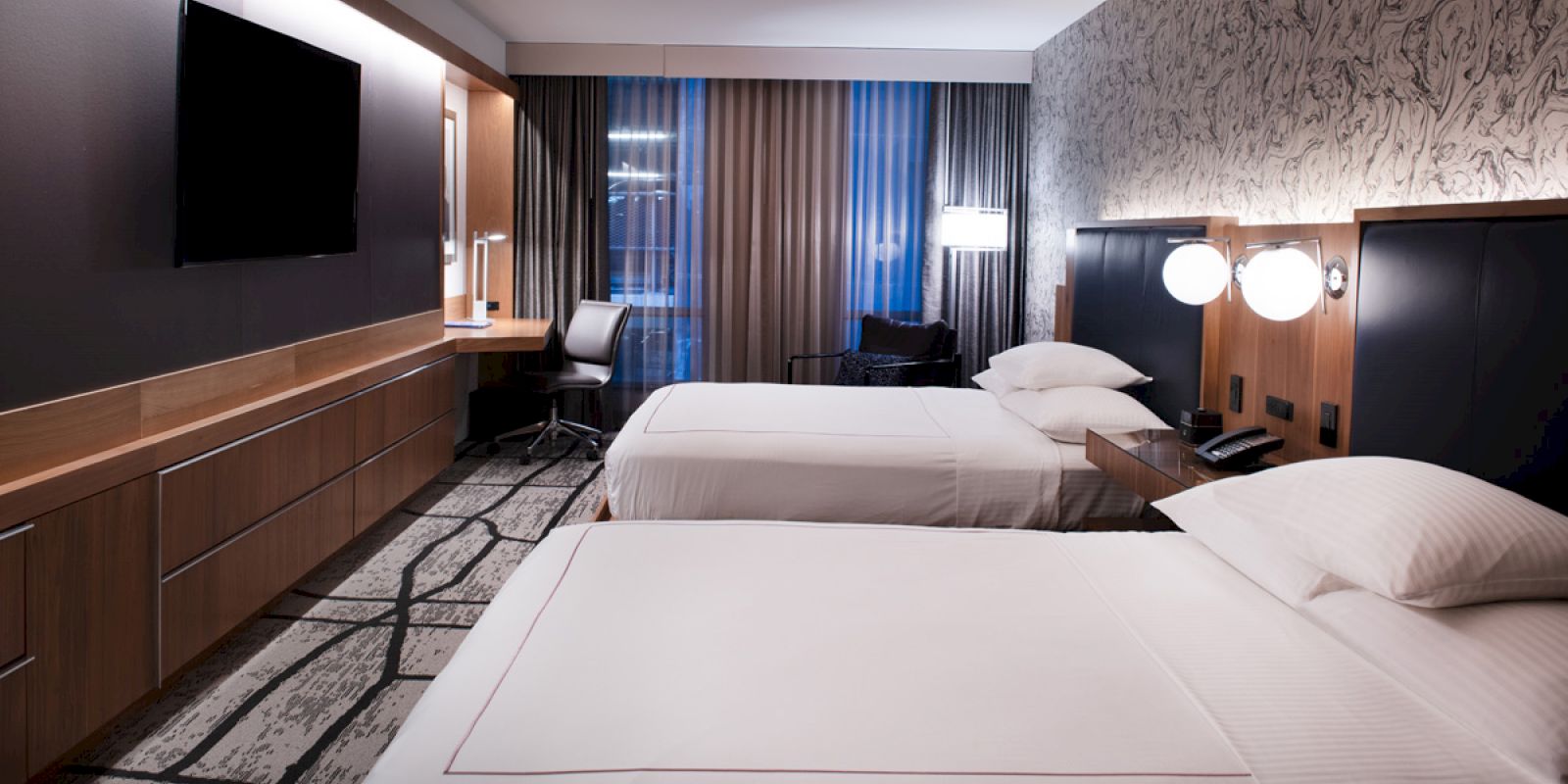 A modern hotel room with two beds, a large TV, a desk and chair, and stylish decor with patterned wallpaper and curtains, softly lit.