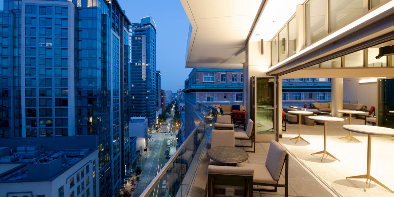 The image shows a modern cityscape with tall buildings and a spacious balcony featuring several chairs and tables overlooking the street below.