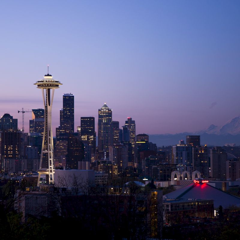 This image depicts the Seattle skyline at dusk, featuring the Space Needle prominently, with Mount Rainier visible in the background.