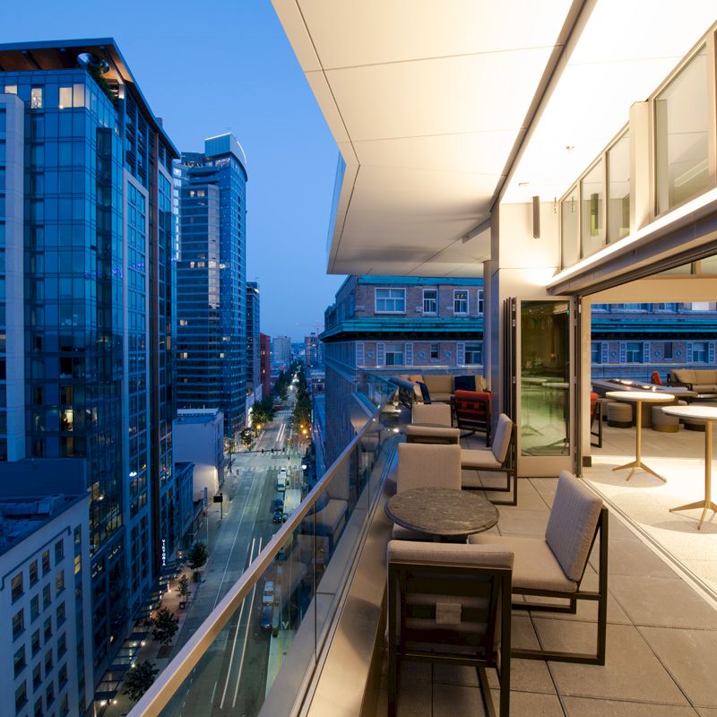A modern cityscape with tall buildings is viewed from a rooftop terrace in the evening, featuring outdoor seating and tables.