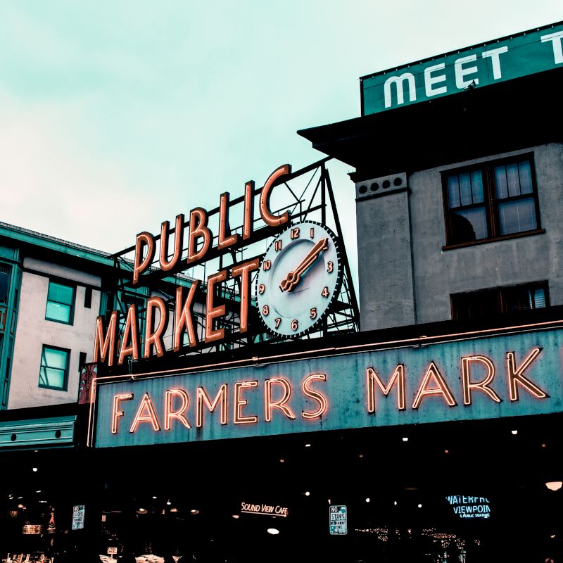 The image shows the iconic neon sign of "Public Market" and "Farmers Market" at Pike Place Market in Seattle, with surrounding buildings and a clock.