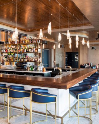 This image shows a modern bar with barstools, hanging lights, and a well-stocked bar counter. In the background, there is a lounge area with seating.