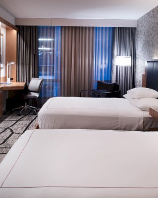 A modern hotel room features two double beds, a TV, a desk with a chair, and wall lamps, with large windows and patterned wallpaper.