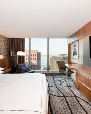 A modern hotel room with a large bed, flat-screen TV, desk, chair, floor-to-ceiling window, and a view of city buildings and the waterfront.