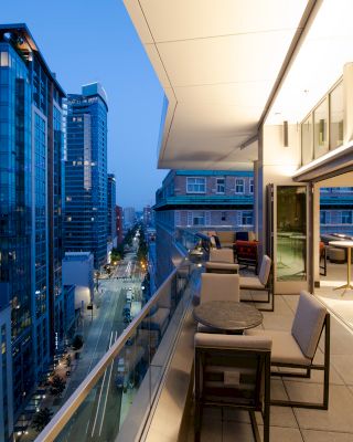 A modern urban balcony with seating, overlooking a cityscape with tall buildings at dusk, featuring lit interiors and a serene ambiance.