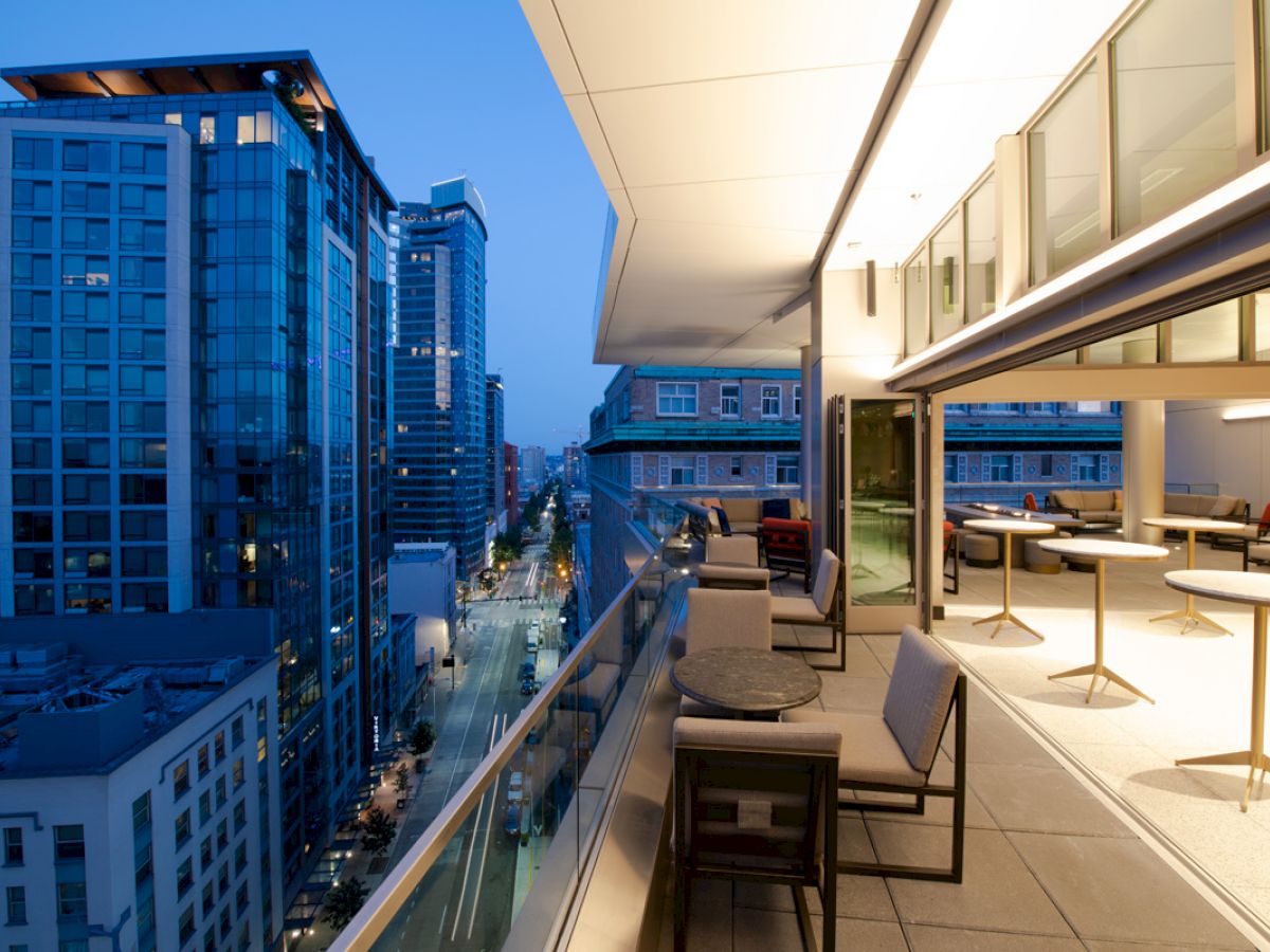 This image shows a modern cityscape at dusk, with tall buildings and a luxurious balcony with seating. The atmosphere is urban and elegant.