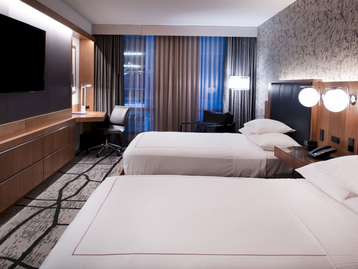 A modern hotel room with two beds, a wall-mounted TV, a desk with a chair, and a large window with curtains. The room appears clean and well-lit.