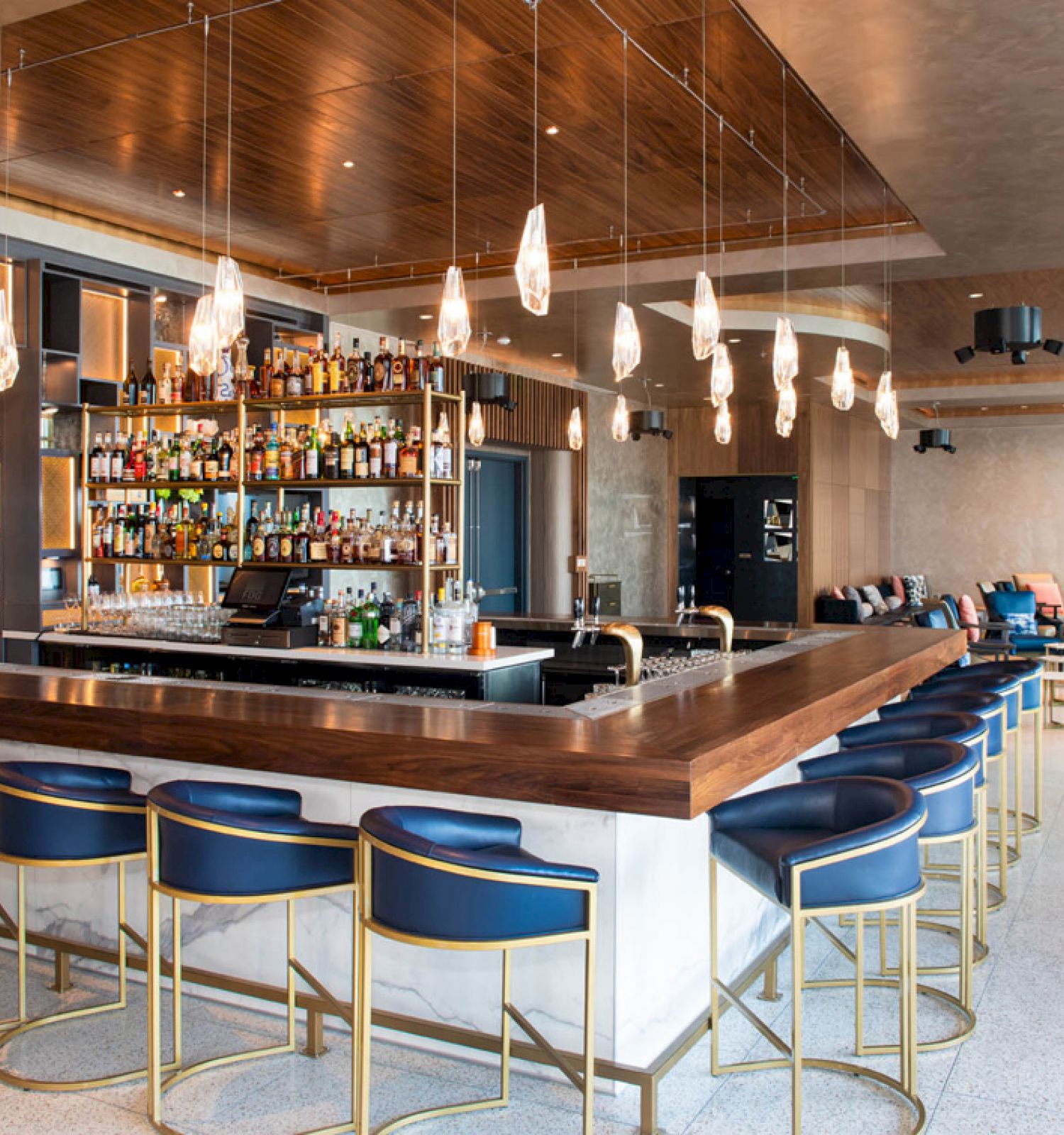 The image shows a modern bar with blue and gold stools, overhead lighting, and a stocked bar. The space is sleek with contemporary decor.