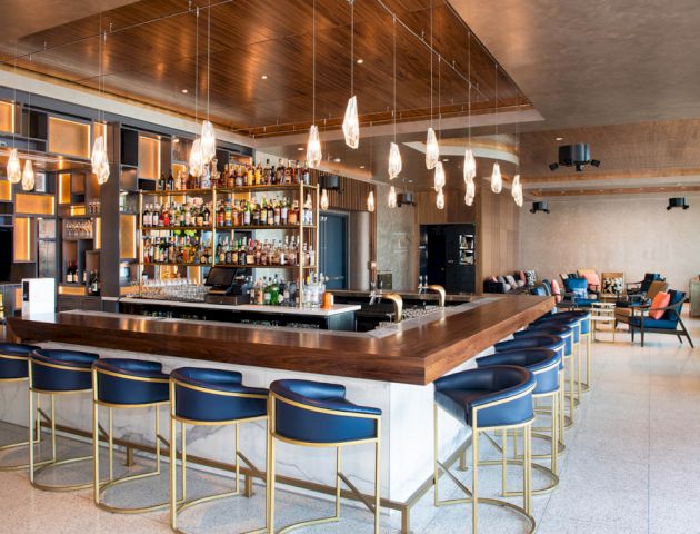 A modern bar with stylish seating, hanging lights, and a fully stocked liquor display behind the counter, set in a spacious, well-lit environment.