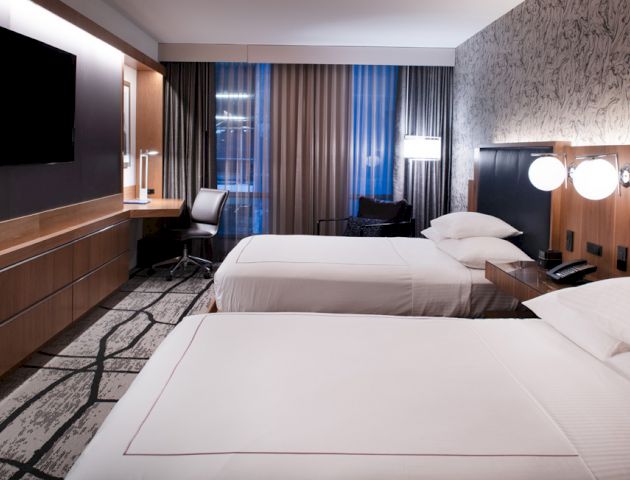 This image shows a modern hotel room with two neatly made beds, a wall-mounted TV, a desk and chair, and stylish lighting fixtures.
