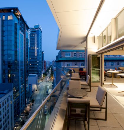 A modern cityscape at dusk features buildings, street lights, and an elegant balcony with seating and tables on the right side.