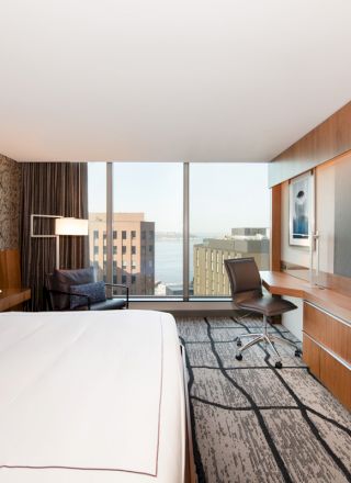 This image shows a modern hotel room with a large bed, flat-screen TV, desk, chair, and window offering a cityscape view.