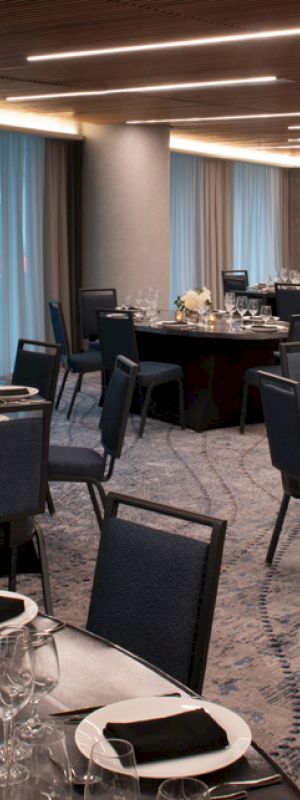 A banquet room is set up with round tables covered in black tablecloths, chairs, cutlery, glassware, and elegant centerpieces, under modern lighting.