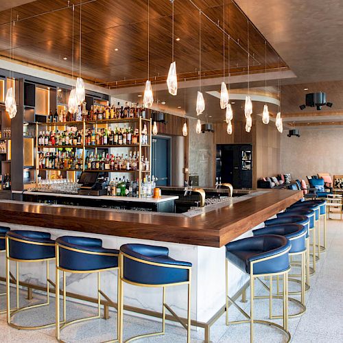 The image depicts a modern bar with a well-stocked liquor shelf, blue barstools, hanging pendant lights, and a sleek, contemporary design.