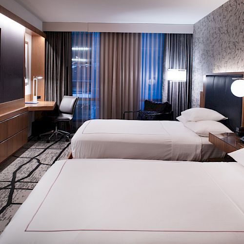 This image shows a modern hotel room with two neatly made beds, a large TV, a desk with a chair, and stylish decor, featuring wall art and good lighting.