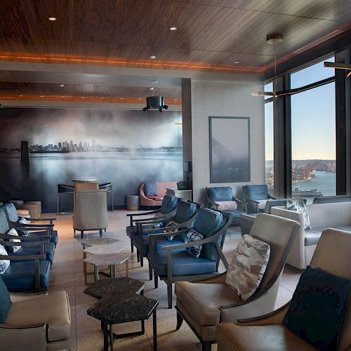 A modern lounge area with large windows, many chairs, and a scenic view of the water and a Ferris wheel outside. The space has a relaxed atmosphere.