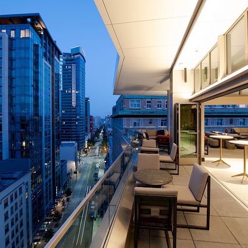 A modern rooftop patio with tables and chairs overlooks a city street lined with tall buildings during twilight.