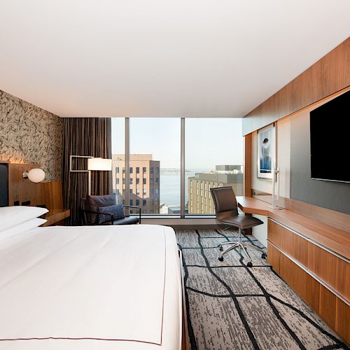 A modern hotel room features a king-sized bed, large TV, desk, armchair, and a window with a cityscape and water view in the background.