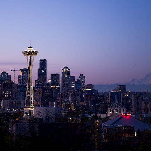 The image depicts the Seattle skyline at dusk, showcasing the iconic Space Needle, various skyscrapers, and a distant mountain range under a twilight sky.
