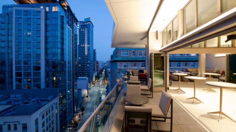 A modern cityscape at dusk featuring tall buildings and a spacious, well-lit balcony with tables and chairs overlooking the street.