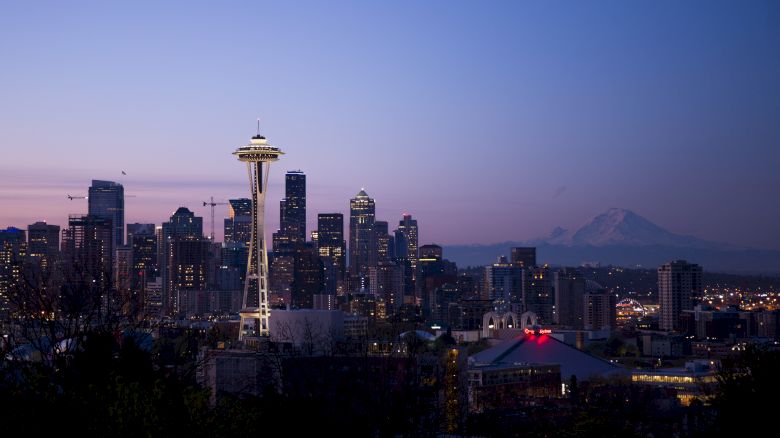The image shows the Seattle skyline at dusk, featuring the Space Needle prominently with Mount Rainier in the background and city buildings illuminated.
