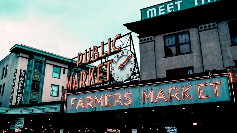 This image shows the iconic signage of a Public Farmers Market in a city, with a clock and neon lights prominently displayed on the buildings.