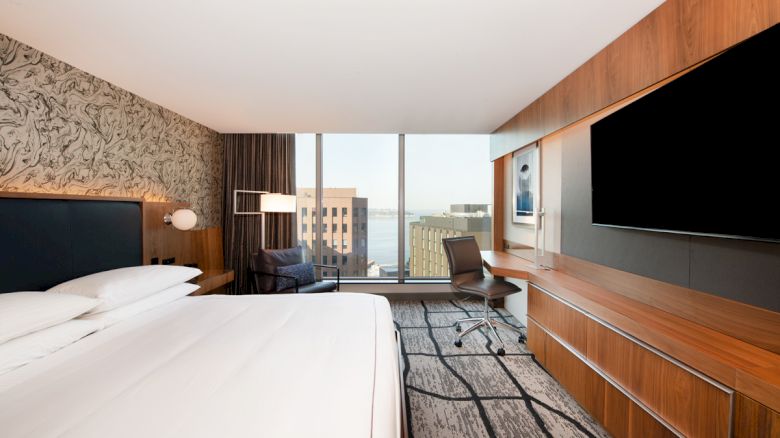A modern hotel room with a large bed, wall-mounted TV, desk, chair, and a view of city buildings through a large window.